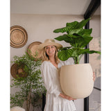Een vrouw in een witte jurk en hoed glimlacht terwijl ze de Kunstplant Calathea UV 72 cm binnenshuis vasthoudt, met op de achtergrond gevlochten manden en nog een plant.