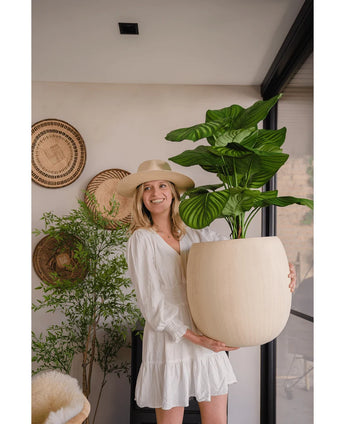 Een vrouw in een witte jurk en hoed glimlacht terwijl ze de Kunstplant Calathea UV 72 cm binnenshuis vasthoudt, met op de achtergrond gevlochten manden en nog een plant.