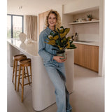 Een glimlachende persoon in spijkerkleding staat in een moderne keuken met in zijn handen een Kunstplant Magnolia in een pot van 46 cm, een onderhoudsvriendelijke plant. De keuken, die perfect is voor interieurdecoratie, is voorzien van houten kasten, een wit kookeiland en twee barkrukken.