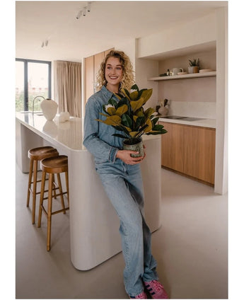 Een glimlachende persoon in spijkerkleding staat in een moderne keuken met in zijn handen een Kunstplant Magnolia in een pot van 46 cm, een onderhoudsvriendelijke plant. De keuken, die perfect is voor interieurdecoratie, is voorzien van houten kasten, een wit kookeiland en twee barkrukken.