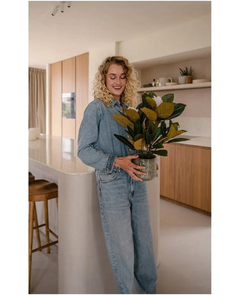Een vrouw met krullend blond haar, gekleed in spijkerkleding, staat glimlachend in een moderne keuken met de Kunstplant Magnolia in een pot van 46 cm in haar handen, omringd door houten kasten en planken – een ideale keuze voor iedereen die op zoek is naar onderhoudsvriendelijke planten om zijn of haar stijlvolle interieur te verfraaien.