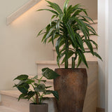 Een hoge Kunstplant Alocasia Cucullata UV 53 cm met hartvormige bladeren in een grote bruine pot met structuur en een kortere plant in een kleine grijze pot staan op een lichte houten trap tegen een neutrale beige muur en leuning.