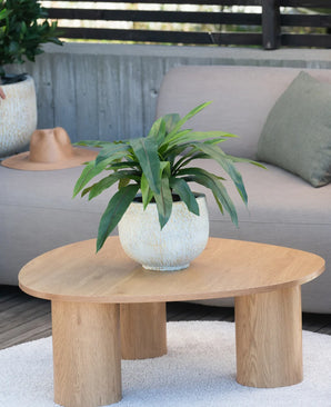 Een Kunstplant Bird’s Deluxe van 60 cm staat op een ronde houten salontafel voor een beige bank, versierd met een groen kussen en een bruin hoedje dat over de armleuning hangt.