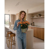 Een vrouw met krullend blond haar, gekleed in een spijkerblouse en een spijkerbroek, staat in een moderne keuken met in haar handen een Kunstplant Daisy UV van 31 cm met feloranje bloemen. Naast haar staan twee houten krukjes bij het keukeneiland.