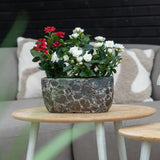 Een Kunstplant Patio-roos 35 cm crème UV staat op een ronde houten tafel voor een beige bank met kussens met een patroon.