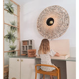 Een jong kind met schouderlang haar zit aan een bureau in een lichte kamer, met de rug naar de kijker gekeerd. Achter haar staan een geweven wandlamp, enkele kasten en de Kunst-hangplant ‘Frosted Varen op bal 76 cm’, genesteld tussen de planten op een houten ladder, naast kleine plankjes met netjes gerangschikte spullen.