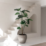 De Kunstplant Monstera 210 cm staat in een minimaal, zonovergoten interieur met witte muren, moderne lijnen, een trap en een beige vloerkleed. Zacht natuurlijk licht benadrukt het strakke ontwerp van de ruimte.
