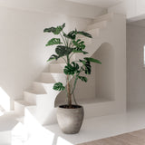De Kunstplant Monstera 210 cm, met brede groene bladeren, staat op een witte vloer in een lichte, minimalistische kamer met witte muren, een trap en natuurlijk zonlicht.