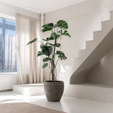 De Kunstplant Monstera 210 cm staat in een pot op een witte vloer naast een moderne trap in een lichte, minimalistische kamer met natuurlijk licht dat door beige gordijnen naar binnen valt.
