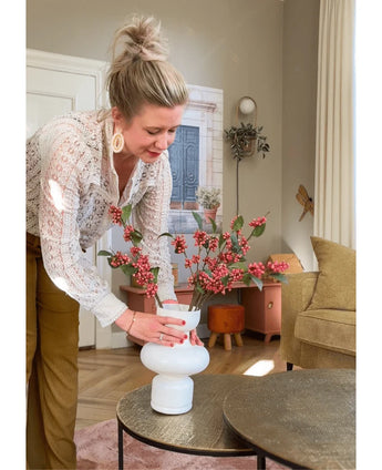 Een vrouw met blond haar in een knot schikt de Kunsttak Berries 64 cm roze in een witte vaas op een ronde tafel in een gezellige woonkamer met aardetinten en een moderne inrichting.