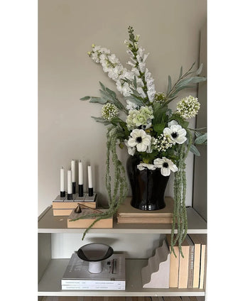 Op een plank tegen een lichte muur staat een zwarte vaas met de Kunstbloem Amaranthus 124 cm groen, naast vijf witte kaarsen, beige boeken en decoratieve voorwerpen in neutrale tinten.