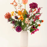 Een witte, geribbelde vaas van keramiek toont het Zijden kunstbloemen boeket Magnus di Lambo - een arrangement van levendige oranje, roze, paarse en gele kunstbloemen met groene bladeren tegen een lichte achtergrond.