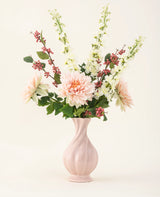 Een roze vaas van keramiek bevat het Zijden kunstbloemen boeket Morning Whisper, met lichtroze dahlia's, witte leeuwenbekjes, groene bladeren en trosjes kleine rode besjes, tegen een effen gebroken witte achtergrond.