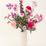 Een vaas met witte structuur bevat het Zijden kunstbloemen boeket Purple Dawn in roze, paarse en lavendeltinten met groen gebladerte, tegen een lichte, neutrale achtergrond.