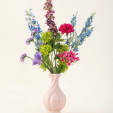 Het Zijden kunstbloemen boeket Blossom Breeze, gerangschikt in een roze vaas van keramiek, heeft levendige blauwe, paarse, rode en groene bloemen - een onderhoudsvrij bloemenboeket tegen een effen lichte achtergrond.