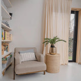 Een gezellig leeshoekje met een beige fauteuil, een rond rieten bijzettafeltje met daarop de Kunstplant Baby Cycas Palm 60 cm, boekenkasten en vitrages die over een glazen deur hangen die uitkomt op de tuin.