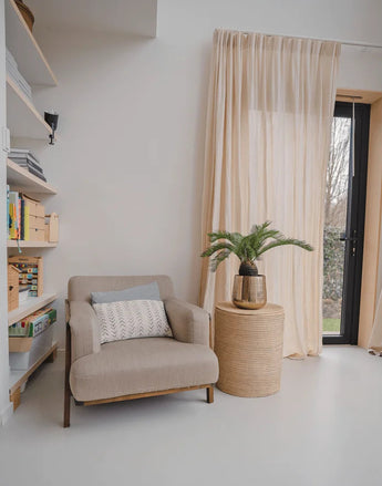 Een gezellig leeshoekje met een beige fauteuil, een rond rieten bijzettafeltje met daarop de Kunstplant Baby Cycas Palm 60 cm, boekenkasten en vitrages die over een glazen deur hangen die uitkomt op de tuin.