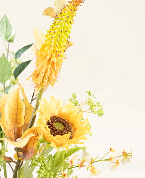 Het Zijden kunstbloemen boeket Amber Light heeft een gele zonnebloem, een lange geeloranje stekelige bloem, groene bladeren en kleine witte bloemen tegen een lichtcrème achtergrond met een amberkleurige gloed.