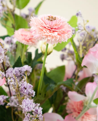 Een close-up van het Zijden kunstbloemen boeket Belle Michelle, met een roze ranonkel, omringd door lichtpaarse en roze bloemen met groene bladeren op de achtergrond.