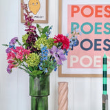 Een groene glazen vaas houdt een Zijden kunstbloemen boeket Blossom Breeze op tafel. Daarachter hangen ingelijste prenten - een met een citroen en "EASY PEASY", een andere met "POESY" - die voor extra charme zorgen. Vlakbij staan een groene kaars en een roze spiraalkaars van Blossom Breeze.