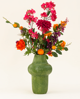 Het Zijden kunstbloemen boeket Fiesta Sunset is gerangschikt in een groene vaas met textuur en toont roze, oranje en paarse bloemen met groene bladeren en kleine oranje vruchten tegen een lichte achtergrond.