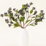 Een Zijden kunstbloemen boeket Japanse Berry Staalblauw met lange stelen, donkerpaarse bessen en staalbladeren staat in een vaas met witte textuur tegen een effen lichte achtergrond.