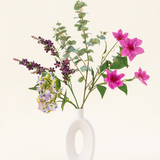 Het Zijden kunstbloemen boeket Lavender Skies, gerangschikt in een moderne witte vaas, bevat roze en paarse kunstbloemen, gele accenten en weelderig groen gebladerte - prachtig tegen een effen lichte achtergrond.