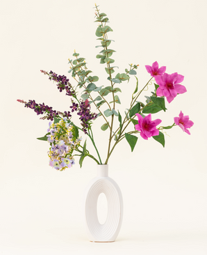 Het Zijden kunstbloemen boeket Lavender Skies, gerangschikt in een moderne witte vaas, bevat roze en paarse kunstbloemen, gele accenten en weelderig groen gebladerte - prachtig tegen een effen lichte achtergrond.