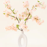 Een witte keramieken vaas met een brede ovale opening toont verschillende groenbladige Zijden kunstbloemen boeket Kersenbloesem lichtroze takken tegen een eenvoudige lichte achtergrond.