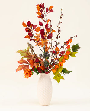 Een vaas met witte structuur toont het Zijden kunstbloemen boeket Chestnut Charm, met herfstbladeren, oranje bloemen en groen gebladerte - een elegant kunstarrangement tegen een effen lichte achtergrond.