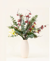 Het Zijden kunstbloemen boeket Midwinter Dream heeft groene bladeren, dennentakken, rode bessen en witte bloemen in een vaas met witte structuur - perfect voor een sereen winterboeket tegen een effen lichte achtergrond.