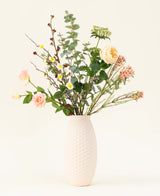 Een vaas met witte structuur toont het Zijden kunstbloemen boeket Blush Blossom, met echte bloemen - lichtroze rozen, gele bloemen en groen gebladerte - tegen een effen lichte achtergrond.