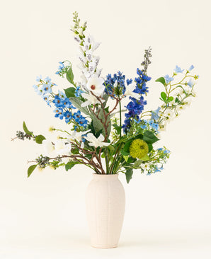 Het Zijden kunstbloemen boeket Cool Breeze, met witte lelies, blauwe ridderspoor en groen gebladerte in een crèmekleurige vaas, steekt af tegen een effen achtergrond voor een verfrissende touch.