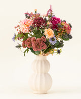In een witte, gebogen vaas van keramiek staat het Zijden kunstbloemen boeket Pretty Pastel - een kleurrijke mix van duurzame bloemen zoals roze rozen, dahlia's, paarse en oranje bloemen en groen - prachtig gerangschikt tegen een lichte achtergrond.