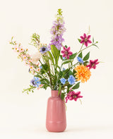 Het Zijden kunstbloemen boeket Bloom & Glow heeft roze, paarse, blauwe, oranje, witte en gele bloemen met groene bladeren in een roze vaas van keramiek tegen een lichte achtergrond.