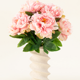 Het Zijden kunstbloemen boeket Peony Soft Pink heeft grote zachtroze pioenrozen en groene bladeren in een golvende, crèmekleurige vaas van keramiek, prachtig weergegeven tegen een effen lichte achtergrond.