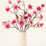De Zijden kunstbloemen boeket Magnolia roze is gerangschikt in een golvende vaas van wit keramiek met groene knoppen, waardoor een elegante display ontstaat tegen een lichte achtergrond.