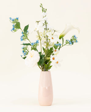 De lichtroze vaas bevat het Zijden kunstbloemen boeket Whispers of White, met witte bloemen, blauwe bloesems en groen gebladerte tegen een effen, lichte achtergrond.