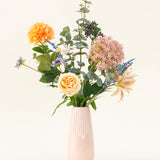 Het Zijden kunstbloemen boeket Earth & Petal is een pastelroze vaas met levensechte zijden bloemen in oranje, roze en geel, plus groen en paarse accenten, afgebeeld tegen een lichte achtergrond.
