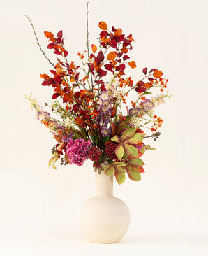 Het Zijden kunstbloemen boeket Golden Autumn heeft een kunstzinnige mix van herfstbladeren en rode, oranje en paarse bloemen in een witte keramieken vaas, tegen een lichte achtergrond.
