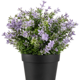 De Kunstplant Buxus paars UV 22 cm heeft een zwarte bloempot met een weelderige kunstplant met kleine lichtpaarse bloemen en groene bladeren tegen een effen witte achtergrond.