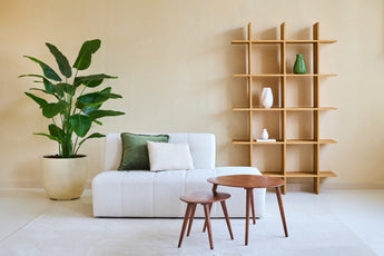 Minimalistische woonkamer met een witte bank, twee kussens, houten nesttafels en een grote Kunstplant Strelitzia Real Touch Deluxe 180 cm voor een tropische sfeer. Een moderne open houten boekenplank met vazen staat tegen de beige muur.