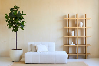 Minimalistische woonkamer met een witte bank, twee kussens en een Kunstplant Ficus Tabaksplant Deluxe 180 cm naast een open houten plank met witte vazen, alles tegen een beige muur en op een licht vloerkleed.