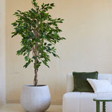 Een Kunstplant Ficus Groen 180 cm in een sierpot staat naast een wit gestoffeerde bank met groene en witte kussens en een klein groen tafeltje, in een minimalistische woonkamer met neutrale muren en een beige vloerkleed.