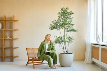 Een vrouw in een groen pak glimlacht terwijl ze op een houten stoel naast de Kunstplant Ruscus 180 cm zit in een lichte, moderne kamer met beige muren, grote ramen en een minimalistische inrichting.