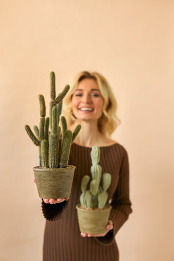 Een glimlachende vrouw in een bruine jurk houdt twee Kunst Cactus Cereus 35 cm planten in pot, een grote en een kleine, tegen een effen beige achtergrond.