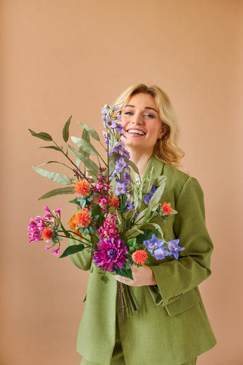 Een lachende blonde persoon in een groen pak houdt het levendige Zijden kunstbloemen boeket Colourful Svenski tegen een effen, lichtbruine achtergrond.