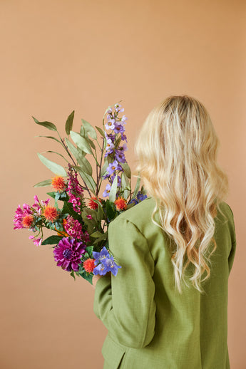 Een persoon met lang, golvend blond haar en een groene jas is van achteren gezien, terwijl hij het Zijden kunstbloemen boeket Colourful Svenski vasthoudt tegen een effen beige achtergrond.