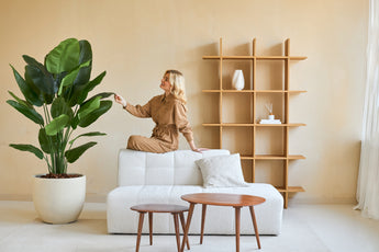 Een vrouw in een bruine jumpsuit zit op de rugleuning van een witte bank en raakt het blad aan van een Kunstplant Strelitzia Real Touch Deluxe 180 cm in een minimalistische woonkamer met houten tafels en planken.