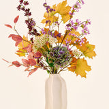 Het boeket ‘Wild Amethyst’ van Zijden kunstbloemen, met herfstbladeren, paarse en witte bloemen en groene hortensia’s, staat in een witte keramische vaas tegen een effen, lichte achtergrond.